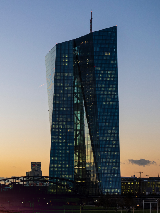A modern skyscraper with a curved glass facade at sunset, with several buildings in the background.