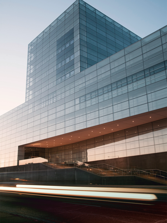 A modern glass building with a blue sky in the background and blurred traffic in the foreground.