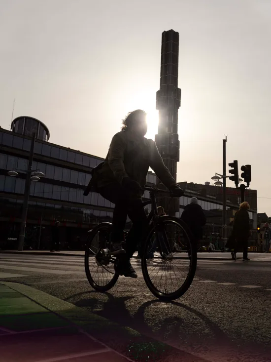 A silhouette of a person on a bicycle in a city street with buildings and a tall tower in the background.