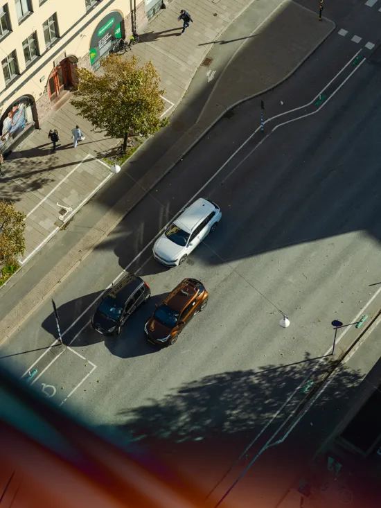 An upper view of a street intersection with cars and pedestrians, surrounded by buildings and trees.