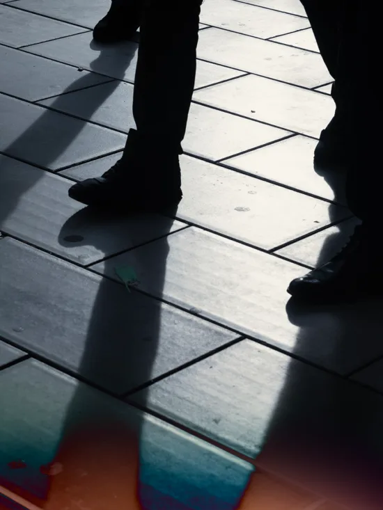 Silhouetted figures casting shadows on a tiled floor, with a colorful light reflection.
