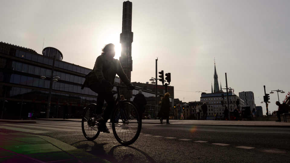 A silhouette of a person on a bicycle in a city street with buildings and a tall tower in the background.