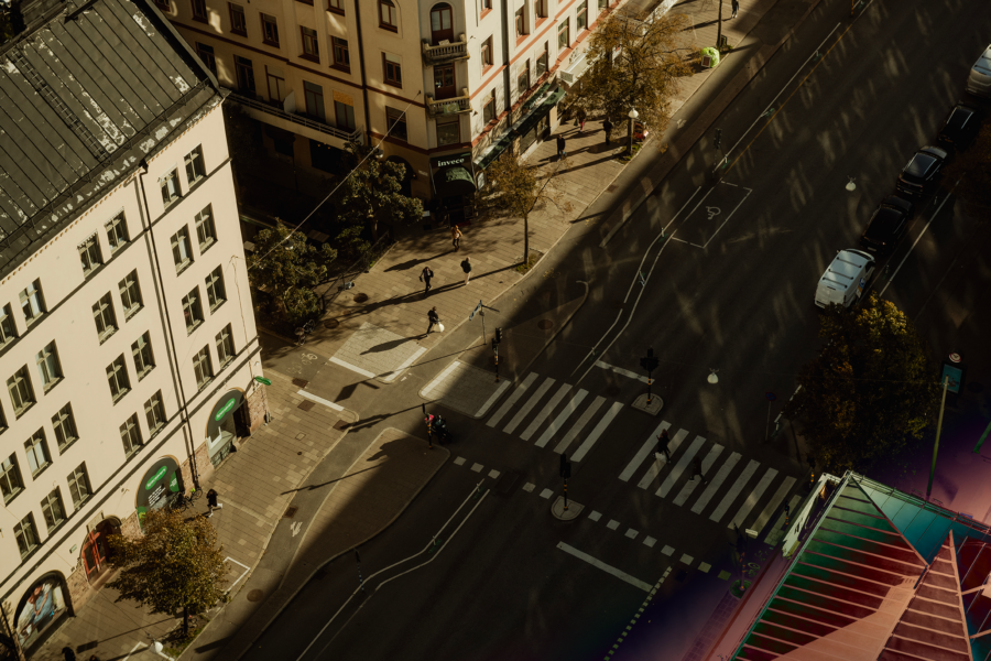 Top-down view of a city street intersection with pedestrians crossing, vehicles, and buildings.