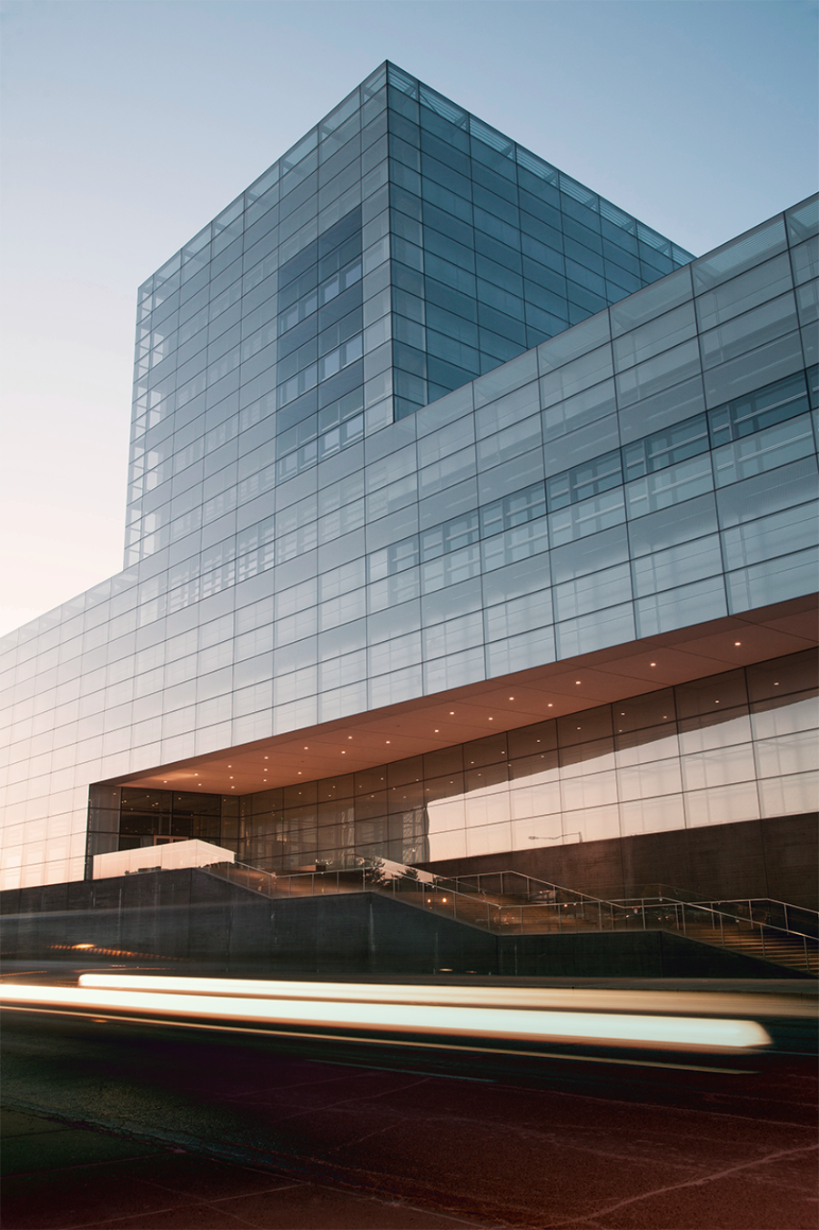 A modern glass building with a blue sky in the background and blurred traffic in the foreground.