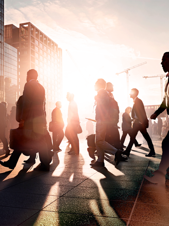 Silhouetted figures walking on a bustling city street at sunset, with tall modern buildings.