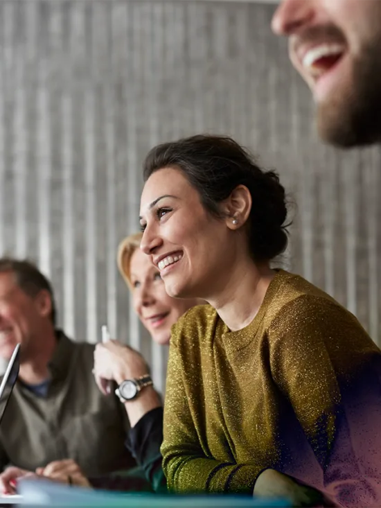 A group of people sitting at a desk, smiling, with a grey background.