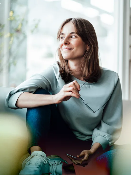 A woman sitting on a couch smiling and looking into the distance, holding a pair of glasses.