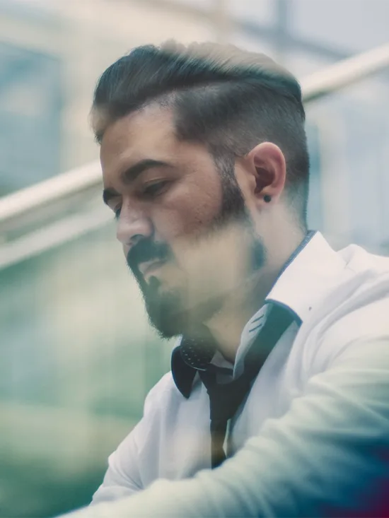 A man with a beard in a white shirt looking down at a phone behind a window.