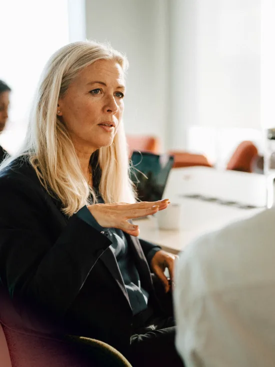 A blonde woman talking to a man at an office table, with colleagues in the background.