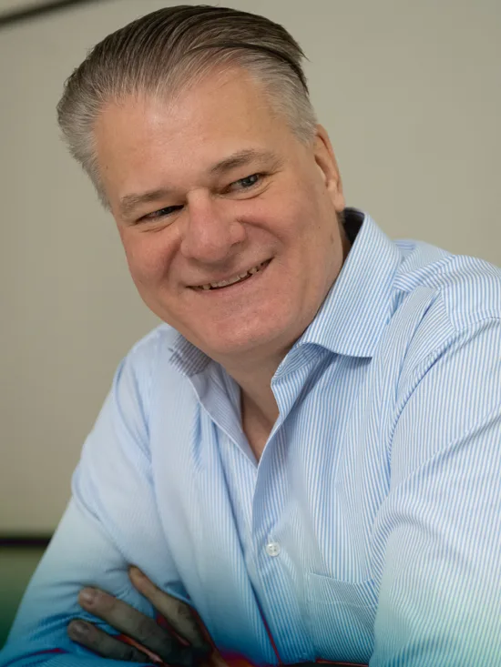 A man with a blue collared shirt sitting in a meeting room, smiling at another person.