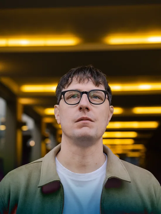 A man wearing a green jacket and glasses, standing in a hallway with yellow lights.
