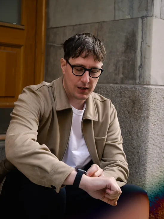 A man with a beige jacket sitting in an entrance, with a stone wall in the background.
