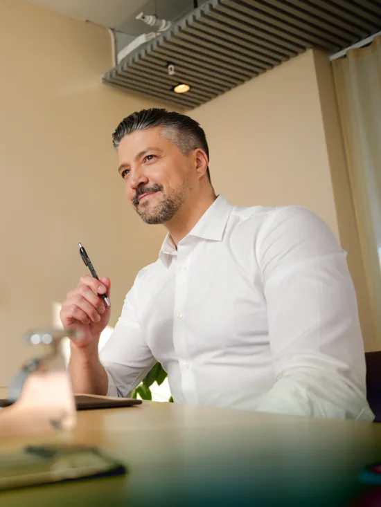 Man in a white shirt sitting at a desk with a pen in his hand, looking into the distance.