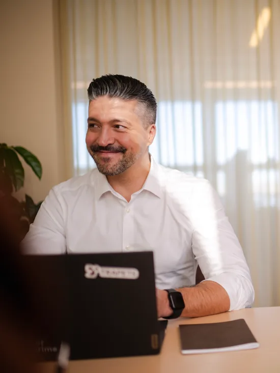 Two men smiling and sitting next to each other at a desk in an office setting.