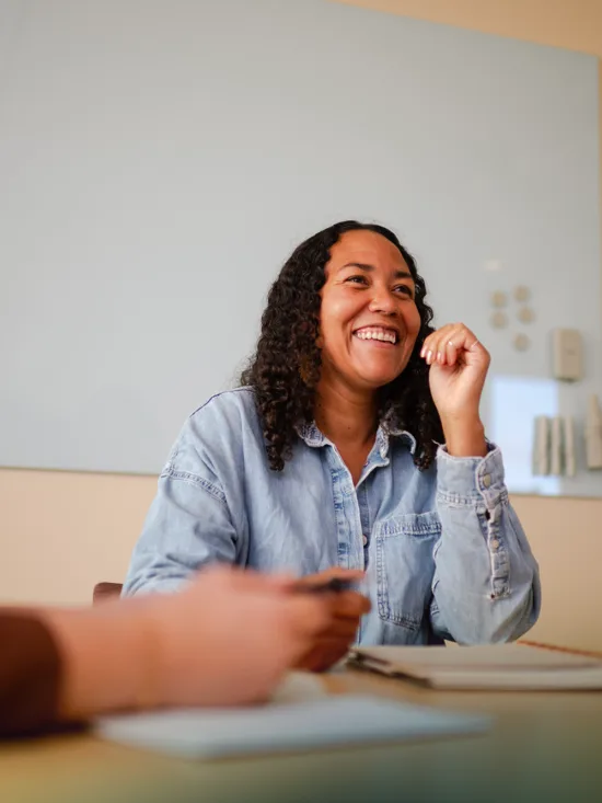 Two laughing women sitting at a table in a meeting room, with a notebook and a laptop.