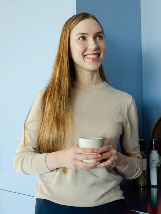 A woman with long hair and a beige sweater smiling and holding a cup of coffee in a blue kitchen.