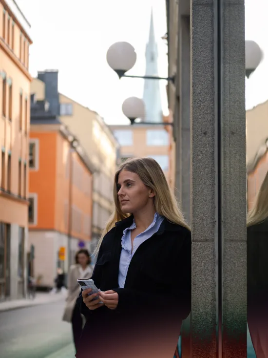 Street scene with buildings and a woman holding a phone, looking in the distance.