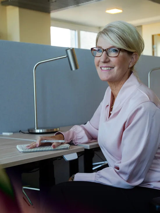 A woman wearing glasses sitting at an office desk next to a computer and a phone.