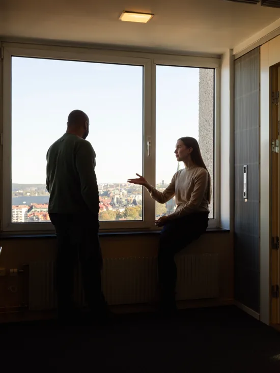 Two people in an office with a view, one standing and one sitting, with a reflection on a glass door.