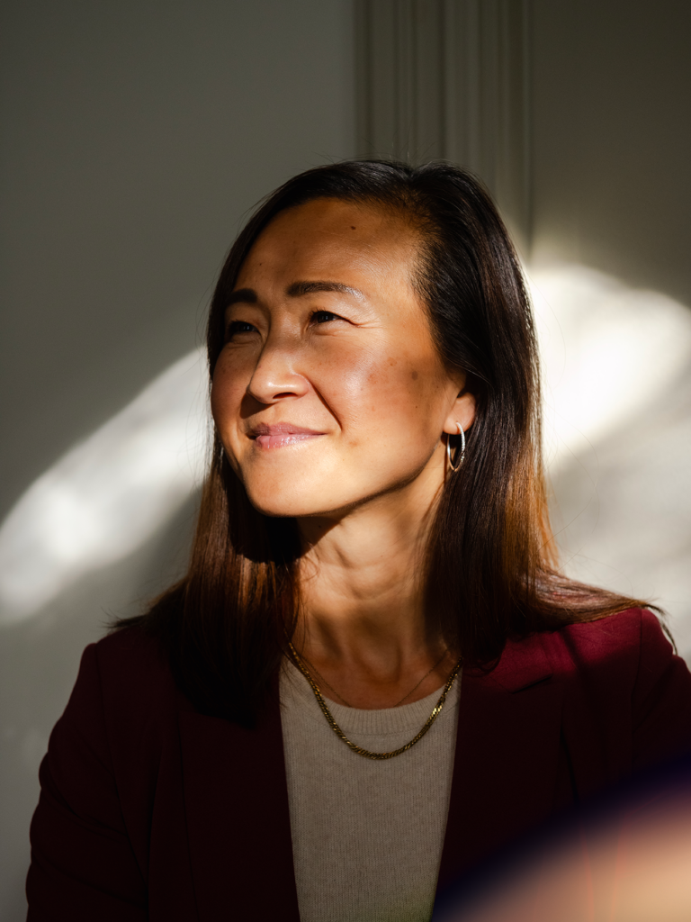 A woman with a red blazer standing by a sunny window lighting up a shadow over her face.