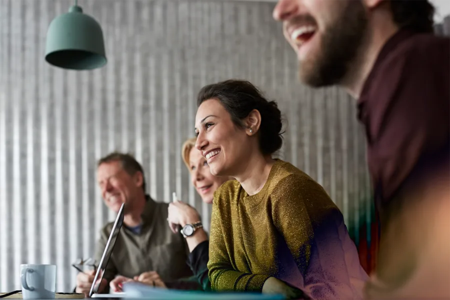A group of people sitting at a desk, smiling, with a grey background.