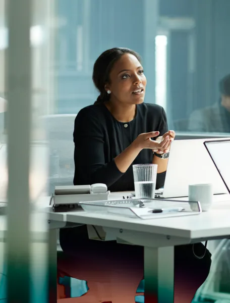 A woman sitting at a desk with office supplies and a colleague in the background.