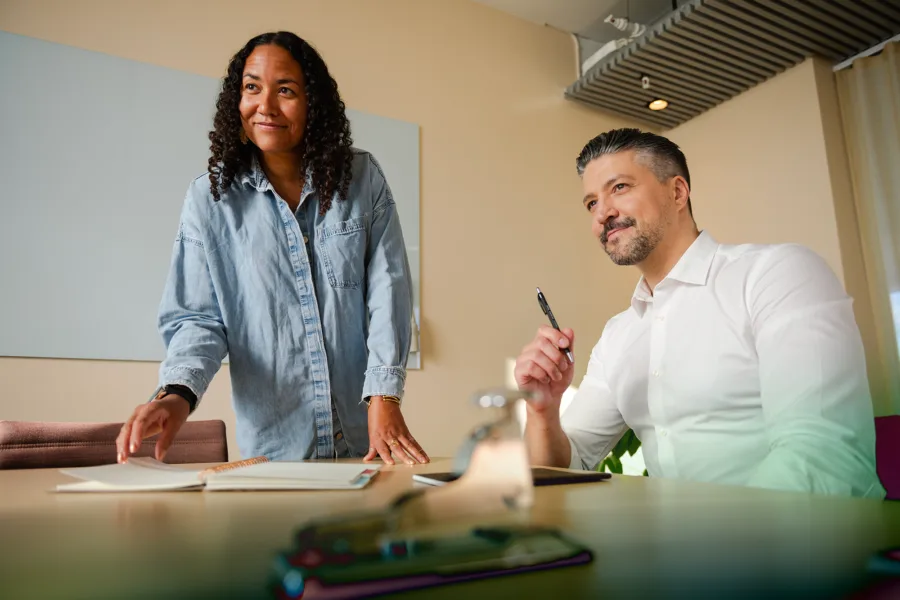 Two people in a modern office, one standing and the other seated, engaged in a discussion.