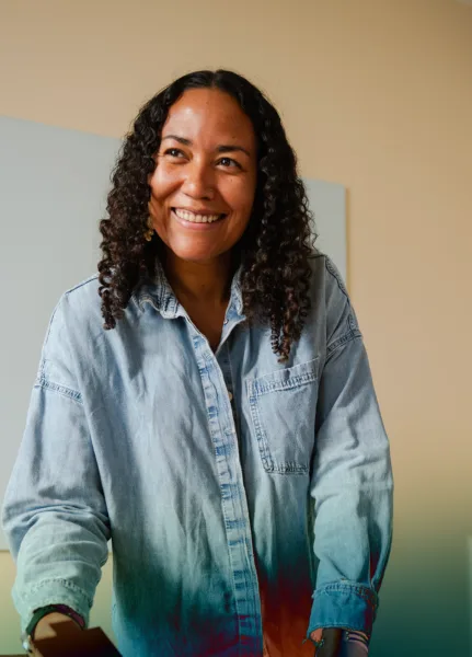 A woman in a blue denim shirt and curly hair smiling and looking into the distance.