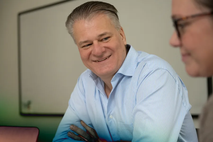 A man with a blue collared shirt sitting in a meeting room, smiling at another person.