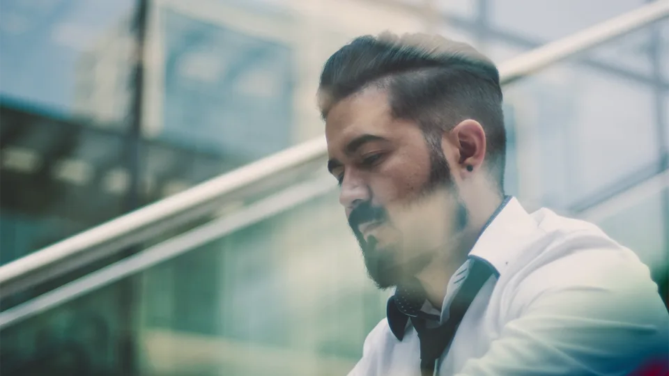 A man with a beard in a white shirt looking down at a phone behind a window.