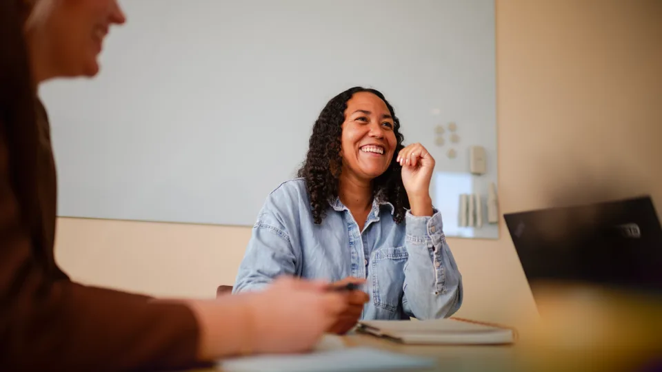 Two laughing women sitting at a table in a meeting room, with a notebook and a laptop.