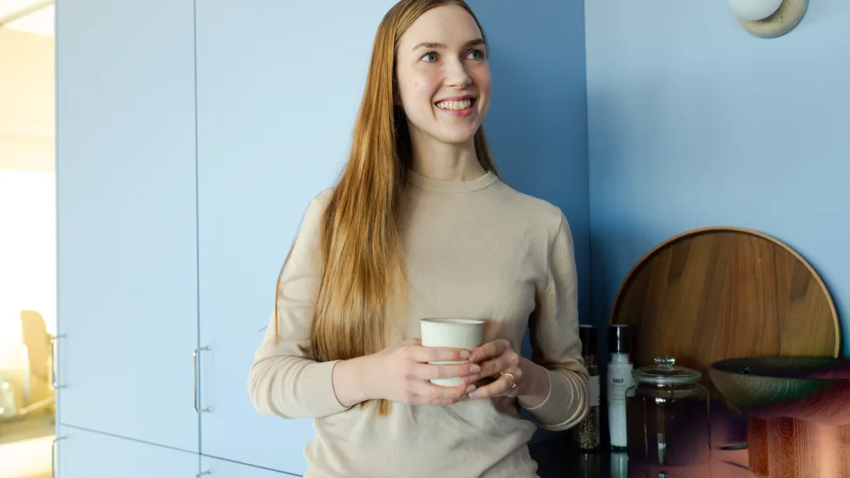 A woman with long hair and a beige sweater smiling and holding a cup of coffee in a blue kitchen.