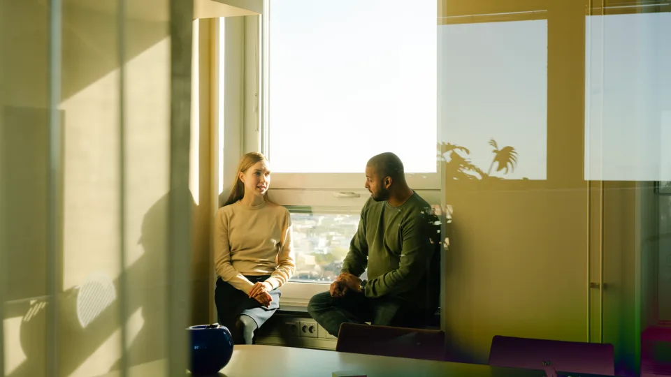 Two people sitting next to a window, in a brightly lit office.