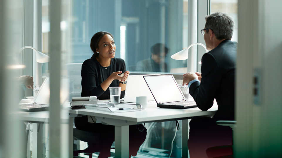 Two people chatting in a modern office, one facing away, at a desk with windows in the background.