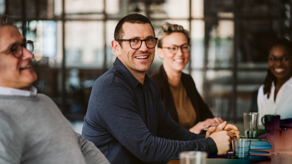 A man in a dark-blue shirt and glasses sitting at an office table, smiling along with three co-workers in the background.