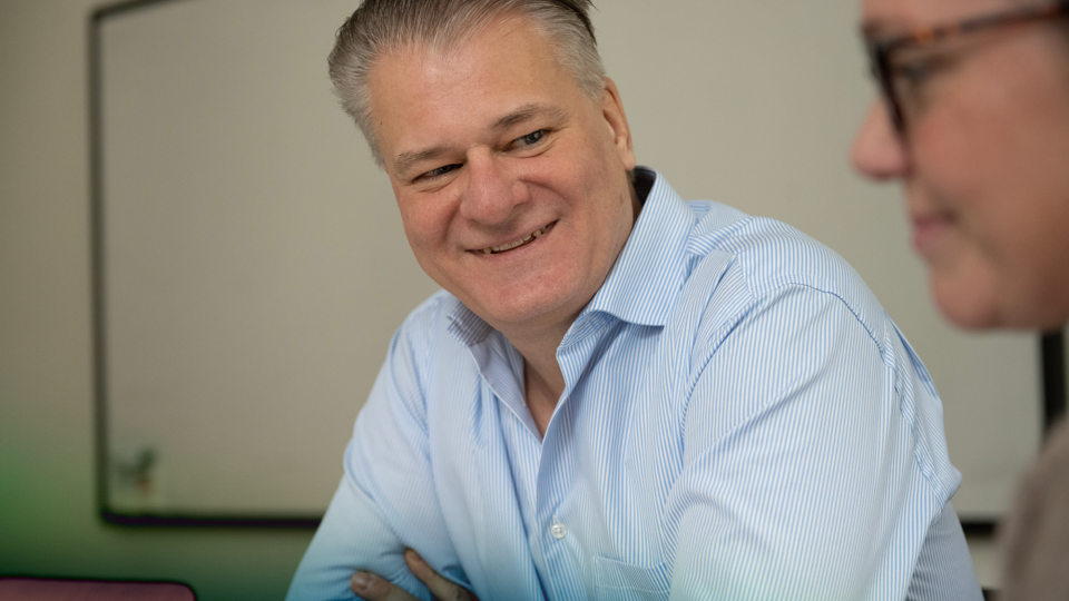 A man with a blue collared shirt sitting in a meeting room, smiling at another person.