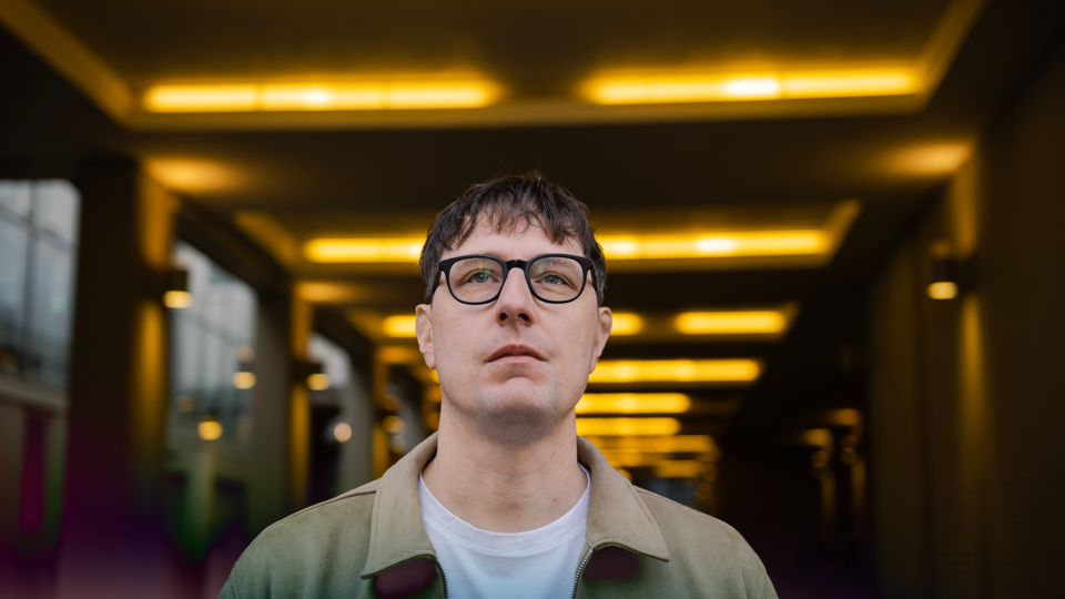 A man wearing a green jacket and glasses, standing in a hallway with yellow lights.