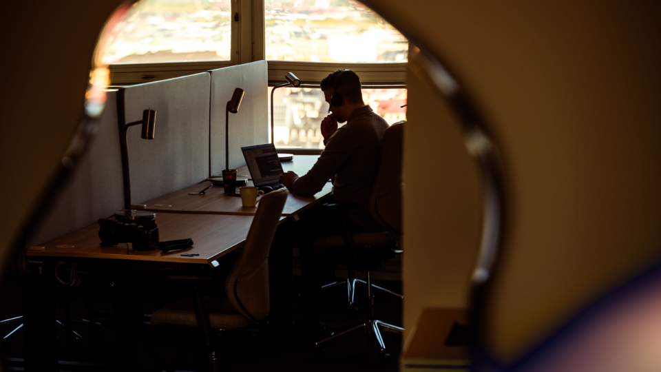 A silhouette of a person working at a desk with a computer and other equipment, seen through a circular window. 