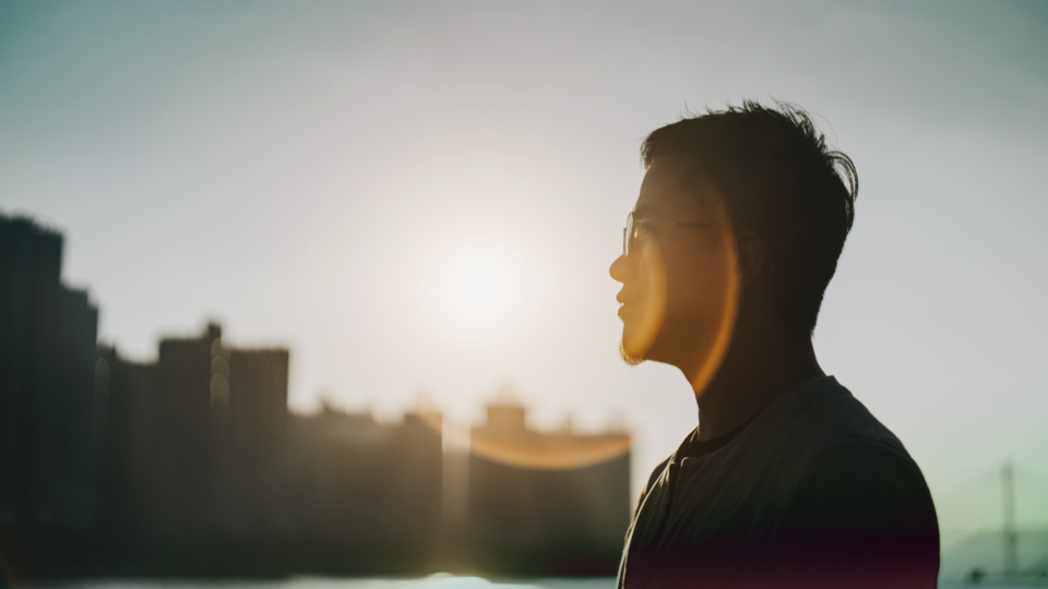 Person with glasses sitting in front of sunset city skyline, casting shadows on his face.