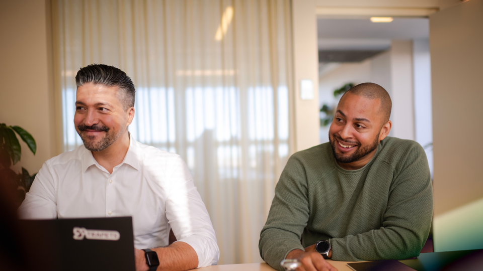 Two men smiling and sitting next to each other at a desk in an office setting.