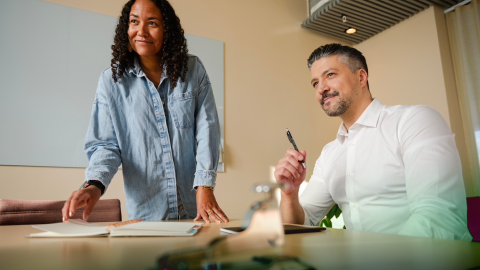 Two people in a modern office, one standing and the other seated, engaged in a discussion.