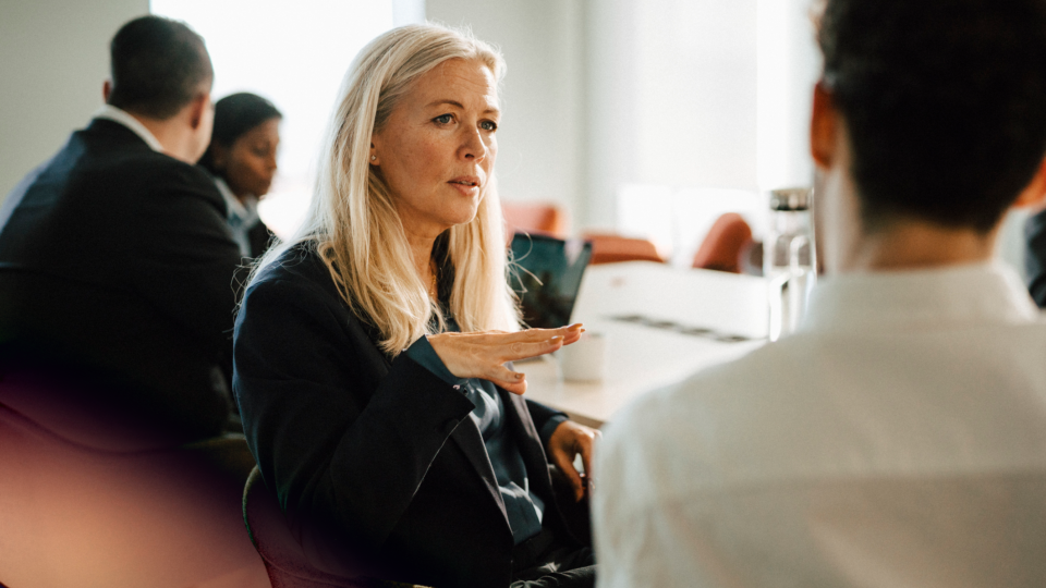 A blonde woman talking to a man at an office table, with colleagues in the background.