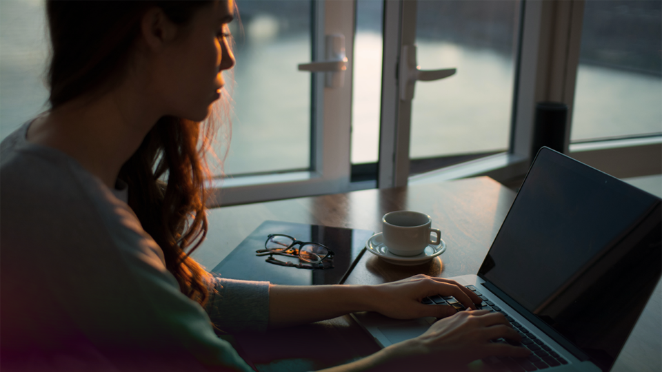 A woman with working on a laptop, with a pair of glasses, a notebook, and a coffee cup beside her.