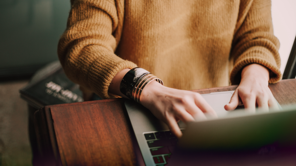 A person in a mustard colored sweater working on a laptop.