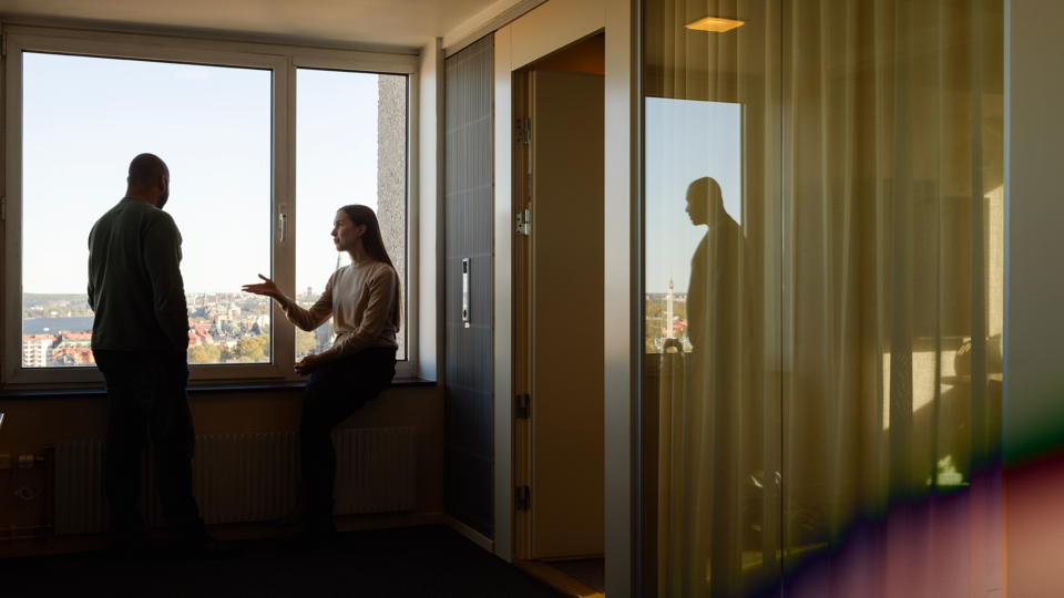 Two people in an office with a view, one standing and one sitting, with a reflection on a glass door.
