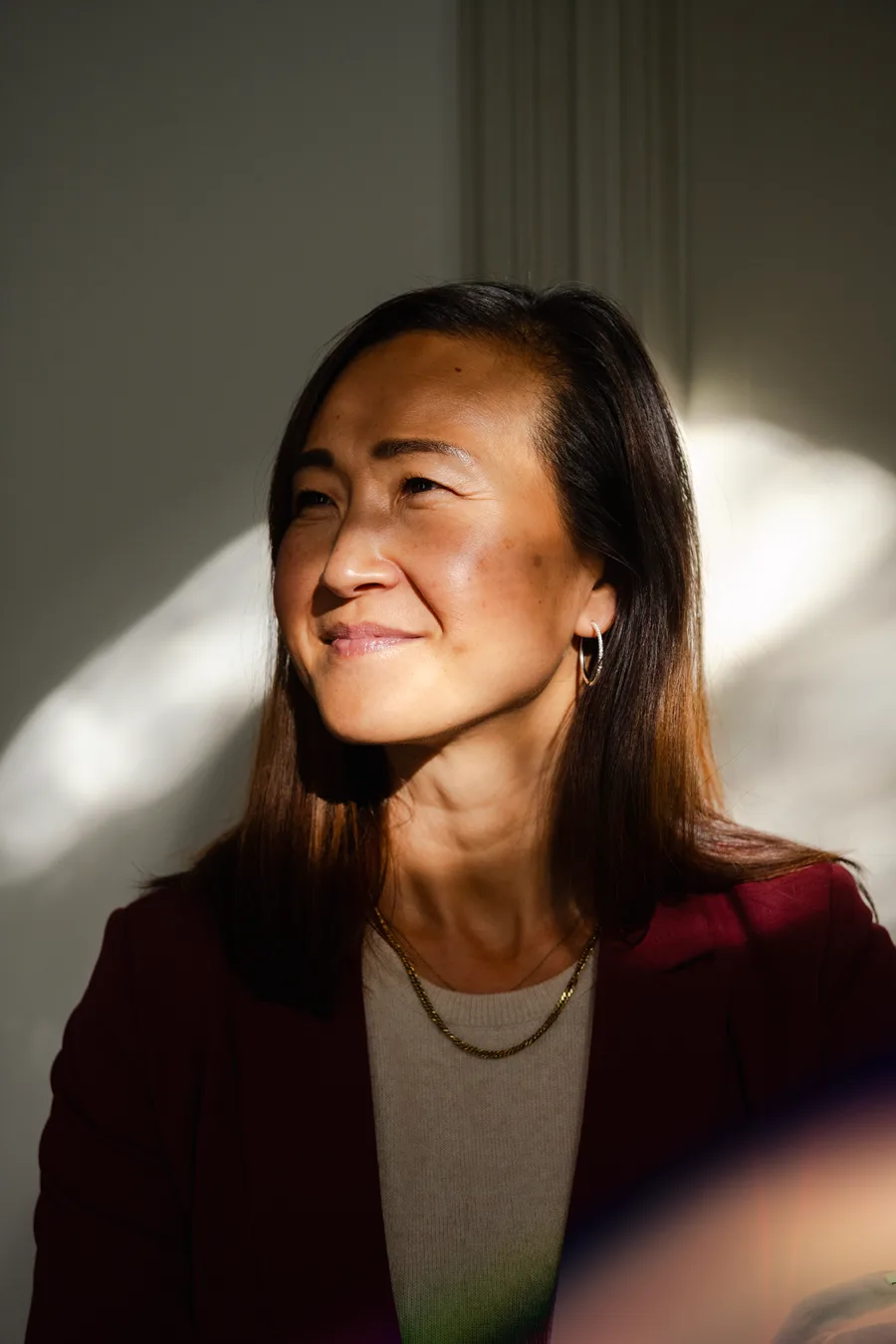 A woman with a red blazer standing by a sunny window lighting up a shadow over her face.