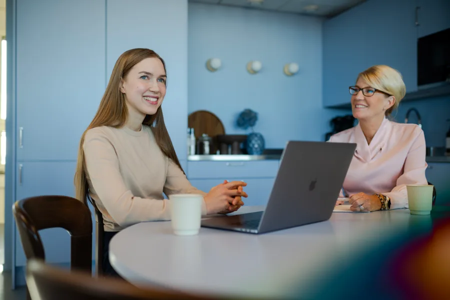 Two women sitting at a kitchen table with a laptop and coffee cups.
