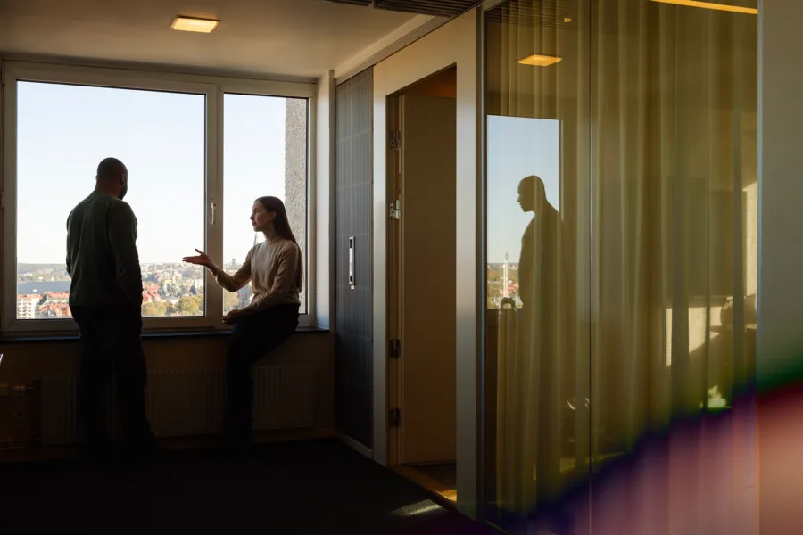 Two people in an office with a view, one standing and one sitting, with a reflection on a glass door.