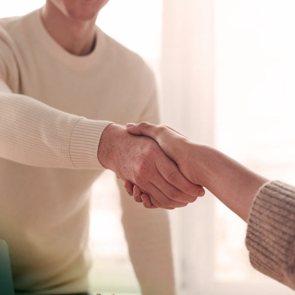 Two people in sweaters shaking hands in a bright room with a window and desk.
