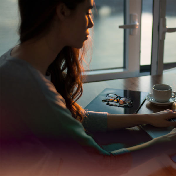 A woman with working on a laptop, with a pair of glasses, a notebook, and a coffee cup beside her.